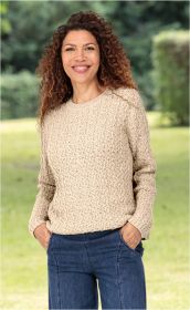 Frau mit lockigem braunem Haar trägt beigen Alpaka-Mix-Pullover mit Rippstrickmuster und blaue Jeans, Hände in den Taschen, lächelt, steht im Grünen, unscharfer Hintergrund aus Bäumen und Gras