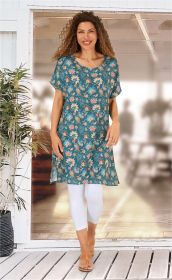Frau trägt blauen Viskose-Kaftan mit floralem Muster, weiße Leggings und braune Sandalen, steht vor einer Glas-Schiebetür mit Pflanzen im Hintergrund, lächelt, lange lockige Haare, helle Wohnraumbeleuchtung