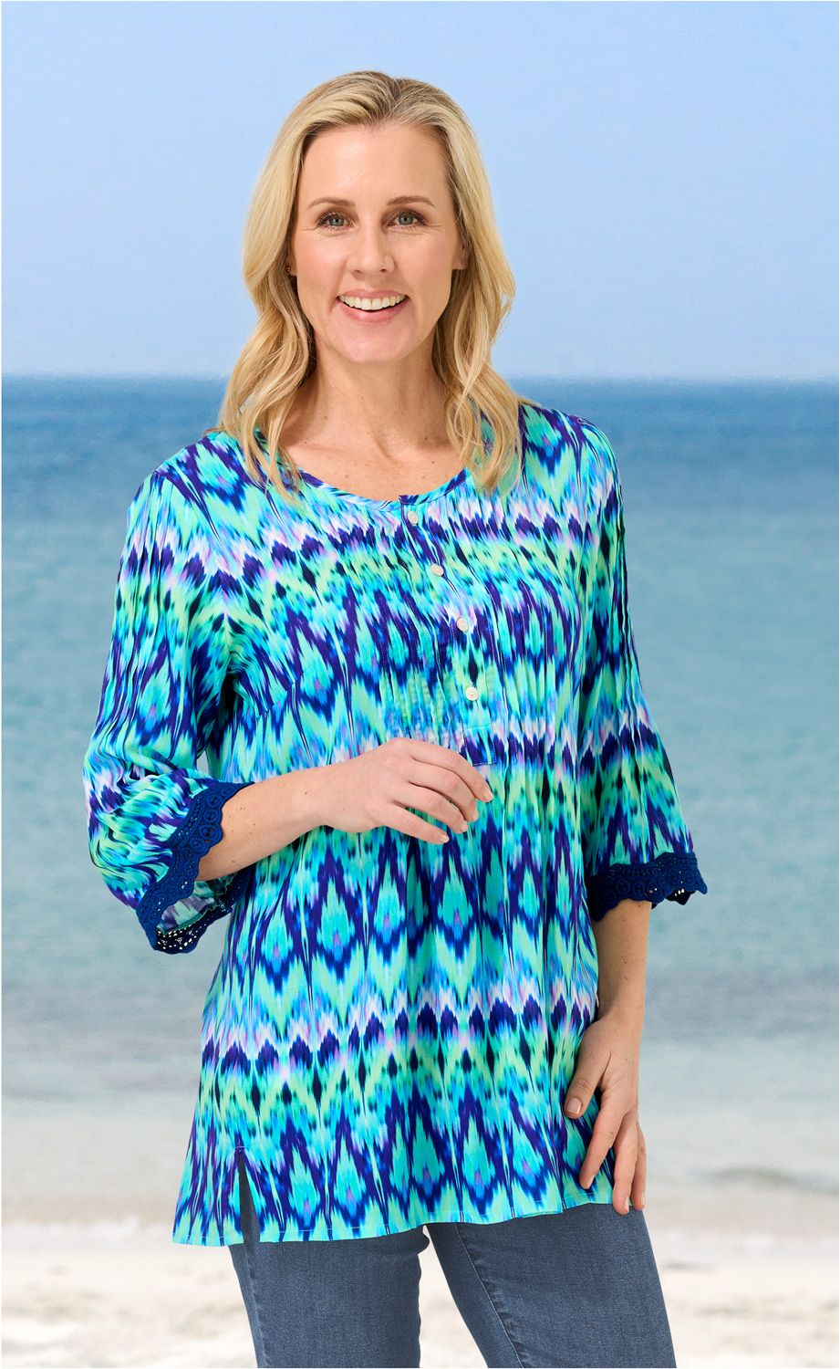 Frau am Strand trägt bunte Modal-Bluse in Blau- und Grüntönen mit 3/4-Ärmeln und Knopfleiste, blaue Jeans, lächelt, hellblonde lange Haare, Meereskulisse im Hintergrund