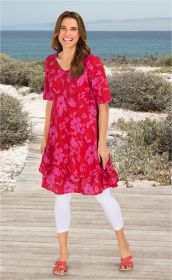 Frau am Strandweg trägt rotes Baumwoll-Mix-Kleid mit pinkem Blumenmuster, weiße Leggings und rote Sandalen, lächelt, braune lange Haare, blauer Himmel und Meer im Hintergrund
