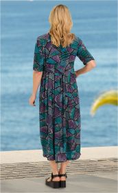 Frau von hinten in einem bunten Viskose-Kleid mit abstraktem Muster in Türkis, Lila und Schwarz, halblange Ärmel, schwarze Sandalen, stehend an einem Wasserufer mit blauem Meer im Hintergrund