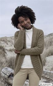 Frau trägt beige Merino-Mix-Strickjacke mit offenem Kragen über weißem Rollkragenpullover, beigefarbene Hose, steht in natürlicher Dünenlandschaft mit trockenem Gras und Treibholz, lächelt leicht, Hand an Kopf gelehnt