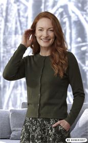 lächelnde Frau mit roten Haaren, grüner Strickjacke und gemustertem Rock, eine Hand an der Seite, in hellem Raum mit grauem Sofa im Hintergrund