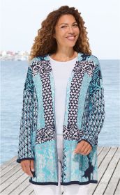 Frau trägt offene Baumwoll-Strickjacke in Blau- und Weißtönen mit floralem und geometrischem Muster, weißes Shirt, weiße Hose, stehend auf Holzsteg am Meer, lockige Haare, lächelnd