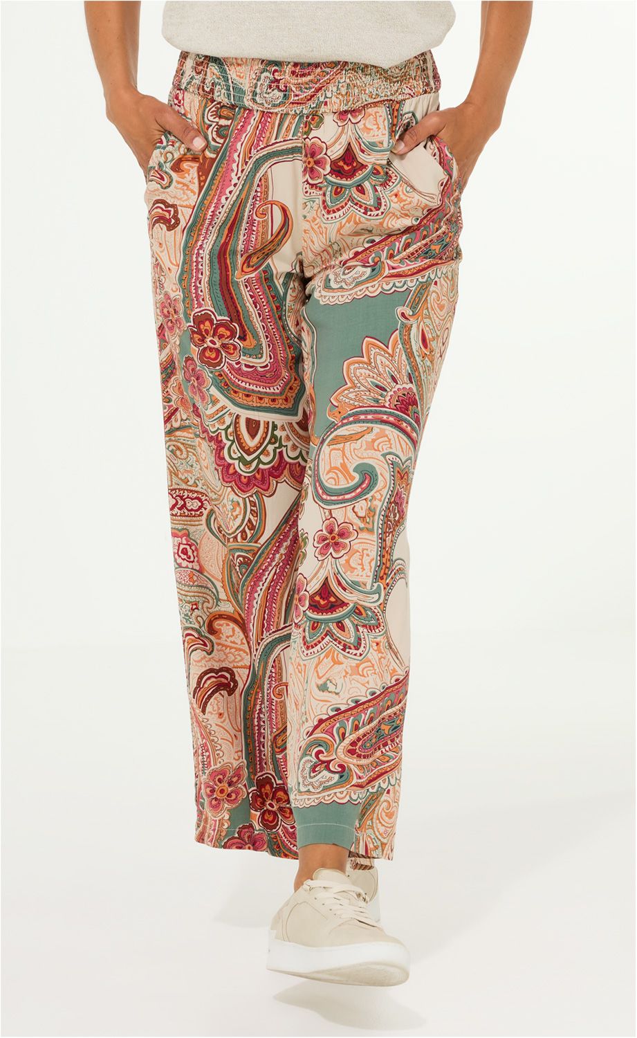 Beinansicht einer locker sitzenden, bunten Modal-Hose mit Paisley- und Blumenmuster in Rot, Grün, Beige, getragen mit beige-weißen Sneakern, Hände in den Taschen, elastischer Bund, leicht verkürzte Beinlänge
