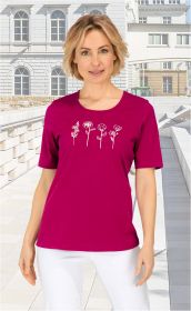 Frau mit kurzem blondem Haar trägt pinkes Baumwoll-Shirt mit weißen Blumenskizzen vorne, kombiniert mit weißen Hosen, vor hellem städtischem Hintergrund mit Treppen und klassischer Architektur