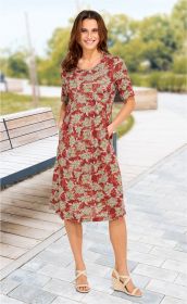 Frau trägt rotes Viskose-Kleid mit floralem Muster in Beige und Grün, halblange Ärmel, steht auf gepflastertem Weg neben Holzbank, beige Keilsandalen, lächelnd, Hände in Taschen, Hintergrund mit Bäumen und Grünflächen unscharf