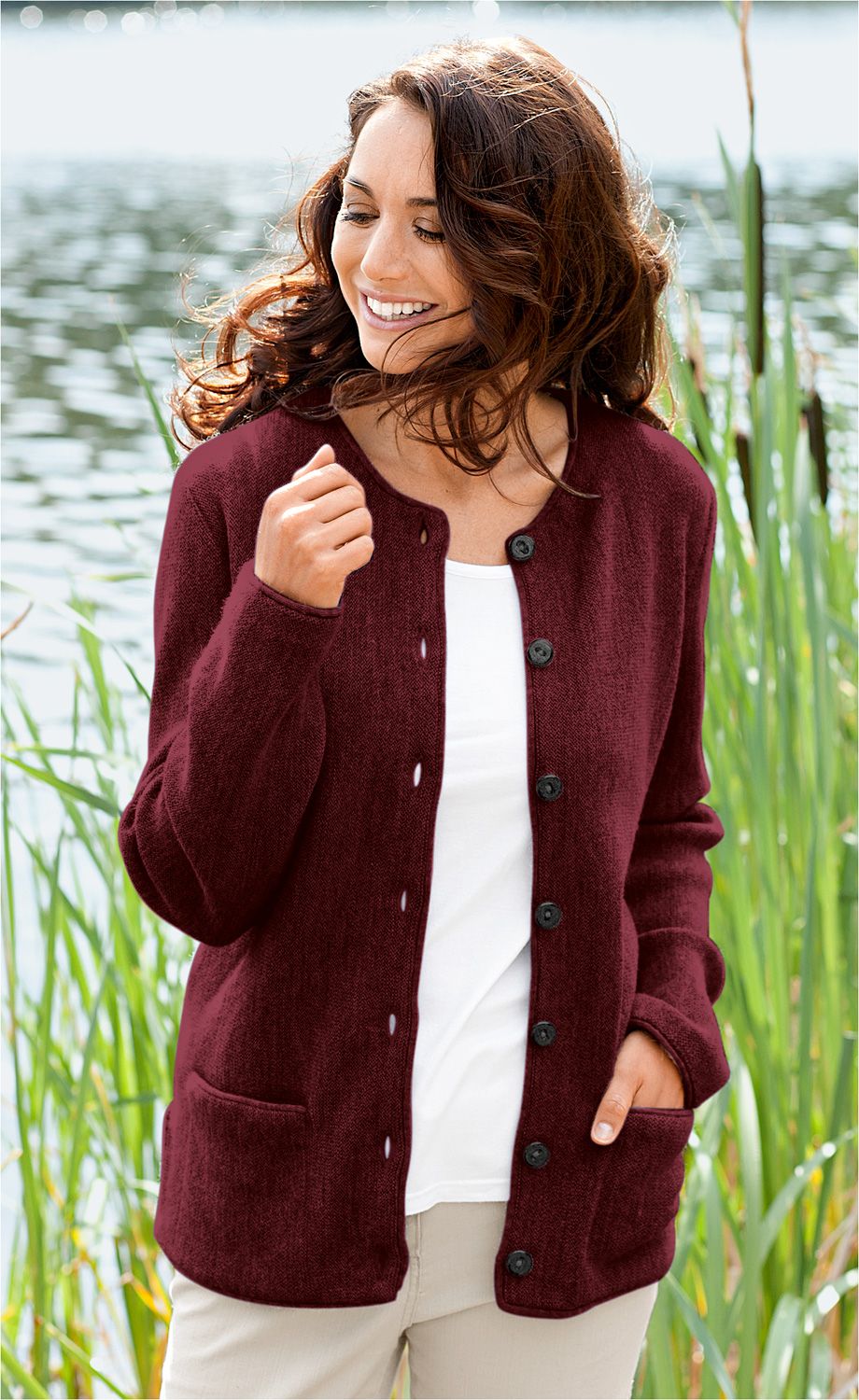 Frau trägt bordeauxrote Alpaka-Jacke mit schwarzen Knöpfen, kombiniert mit weißem Shirt und hellen Hosen, steht lächelnd vor einem Teich mit Schilf im Hintergrund