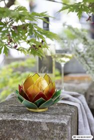 Capiz Windlicht in Lotusblüten-Form mit gelben, roten und grünen gläsernen Blättern auf einem steinernen Gartentisch, umgeben von grünem Laub und einem Weinglas mit Weißwein im unscharfen Hintergrund.