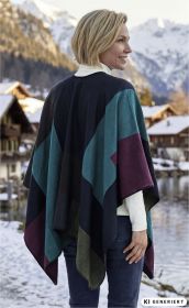 Frau von hinten im farbigen Poncho in Blau, Grün, Lila und Schwarz vor verschneiter Berglandschaft mit See und Holzhütten, trägt weißen Rollkragenpullover und blaue Jeans, Herbstwinterstimmung