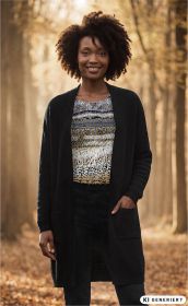 Frau trägt schwarzen Baumwoll-Mix-Strickmantel mit Taschen, darunter gemustertes Shirt in Naturtönen, steht lächelnd im herbstlichen Wald mit Laub auf dem Boden und Bäumen im Hintergrund