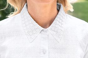 Nahaufnahme weißer Baumwoll-Mix-Bluse mit fein strukturierter Oberfläche, Knopfleiste und spitzem Kragen aus floraler Spitzenstickerei, getragen von Person mit hellem Teint und blonden Haaren, unscharfer grüner Hintergrund