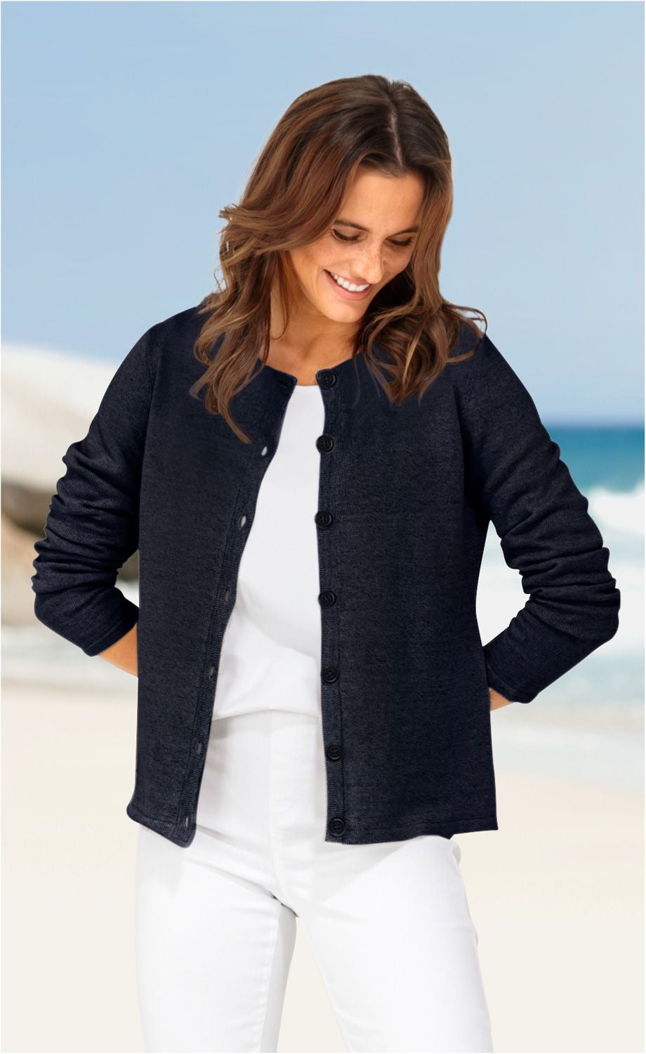 Frau trägt dunkelblaue Leinen-Mix-Strickjacke mit Knöpfen, weiße Bluse und weiße Hose, steht am Strand mit blauem Himmel und Meer im Hintergrund, schaut lächelnd nach unten