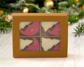 Braunes Kartonpaket mit vier Teelichtern in Form von roten Halbkreisen und weißen Tannenbäumen, dekoriert mit einem verschwommenen Weihnachtsbaum im Hintergrund, auf hellem Holzuntergrund