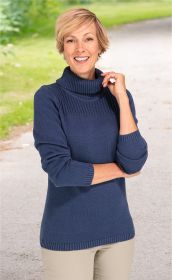 Frau in blauem Baumwoll-Mix-Pullover mit Rollkragen, lächelt, steht auf einem Weg mit Gras im Hintergrund, trägt beige Hose, helle Frisur, rechte Hand berührt Pulloverkragen, linke Hand verschränkt vor dem Körper