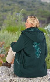 Blonde Frau sitzt auf Stein, trägt dunkelgrüne Baumwoll-Jacke mit Kapuze und türkisfarbenen Spiralstickereien auf Rücken, beige Hose und braune Schuhe, im Hintergrund grüne, bewachsene Natur