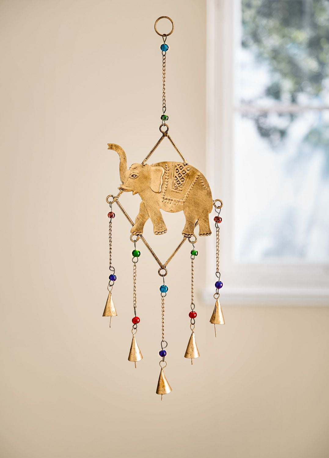 Metall-Mobile 48cm mit einem goldfarbenen Elefanten in der Mitte, verziert mit kleinen bunten Perlen, hängend an einer Kette mit mehreren kleinen Glocken, vor hellem Hintergrund und Fenster mit unscharfen Baumblättern.