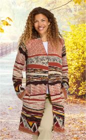 Frau mit lockigen Haaren trägt bunte Baumwoll-Strickjacke mit abstraktem Muster in Rot-, Braun- und Beigetönen, beige Hose, steht im herbstlichen Waldweg mit gelb verfärbten Blättern und heller Beleuchtung.