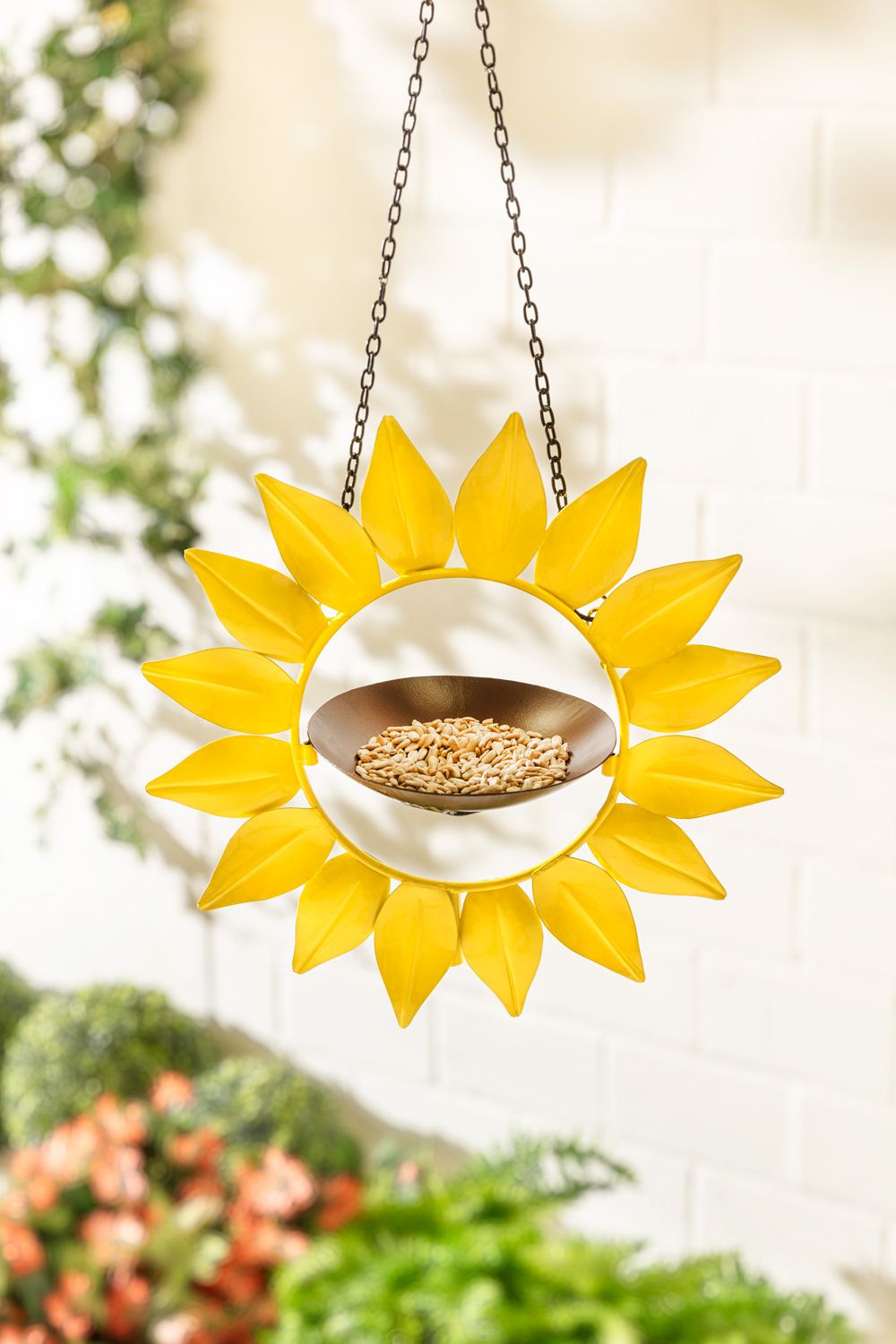 Metall-Futterstation in Sonnenblumenform, gelbe Metallblätter, hängend an Kette, braune Futterschale mit Vogelfutter, unscharfer Hintergrund mit weißen Wand und grünen Pflanzen.