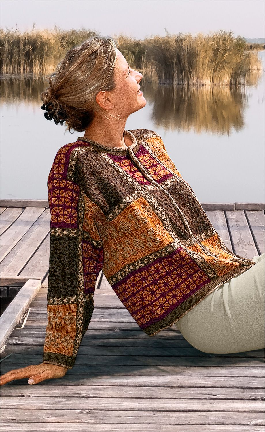 Frau sitzt entspannt auf Holzsteg am See, trägt bunte Alpaka-Strickjacke mit geometrischem Muster in Orange, Braun und Dunkelrot, Haare zum Dutt gebunden, Blick in den Sonnenuntergang gerichtet, Schilf im Hintergrund