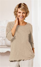 Frau mit kurzem blonden Haar trägt hellbeiges Baumwoll-Shirt mit 3/4-Ärmeln und gewelltem Saum, lächelt, eine Hand am Hals, steht vor hellen Wänden und Möbeln.