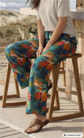 Frau sitzt auf einem Holzstuhl am Strand, trägt bunte Viskose-Hose mit abstraktem Muster in Blau, Orange und Rot, cremefarbenes Kurzarm-Pullover, braune Sandalen, entspannte Sommerstimmung