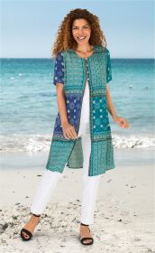 Frau am Strand trägt langes Viskose-Kleid mit buntem geometrischem Muster in Blau- und Grüntönen, dazu weiße Hose und schwarze Sandaletten, lächelnd, lockige rote Haare, blauer Himmel und Meer im Hintergrund