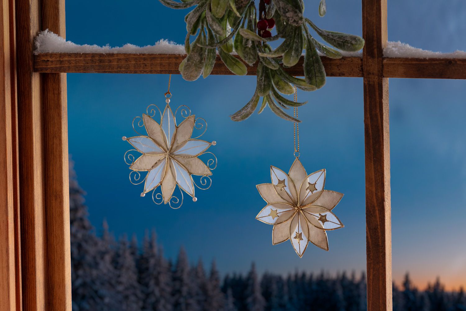 Zwei naturfarbene Capiz-Stern-Ornamente mit goldenen Rändern, hängend an goldenen Schnüren an einem schneebedeckten Holzfensterrahmen, Hintergrund schneebedeckte Bäume und Abendhimmel, oben Mistelzweig mit roten Beeren.