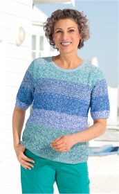 Frau mit kurzen lockigen Haaren trägt einen mehrfarbigen, gestrickten Baumwoll-Pullover in Blau- und Türkistönen mit kurzen Ärmeln, lächelt, steht vor weißer Hauswand und blauem Himmel im Hintergrund