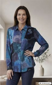 Frau trägt blau-schwarze Modal-Bluse mit Patchwork-Design und floralem Muster, langärmlig, vor neutralem Hintergrund, Hand in Hüfte, kombiniert mit dunkelblauer Jeans, Pflanzenvase im Hintergrund