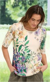 Frau trägt cremefarbenes Baumwoll-Shirt mit buntem Blumen- und Blätterdruck, Halbarm, Rundhalsausschnitt, Hände in olivgrüner Hose in den Taschen, stehend vor unscharf-grünem Hintergrund