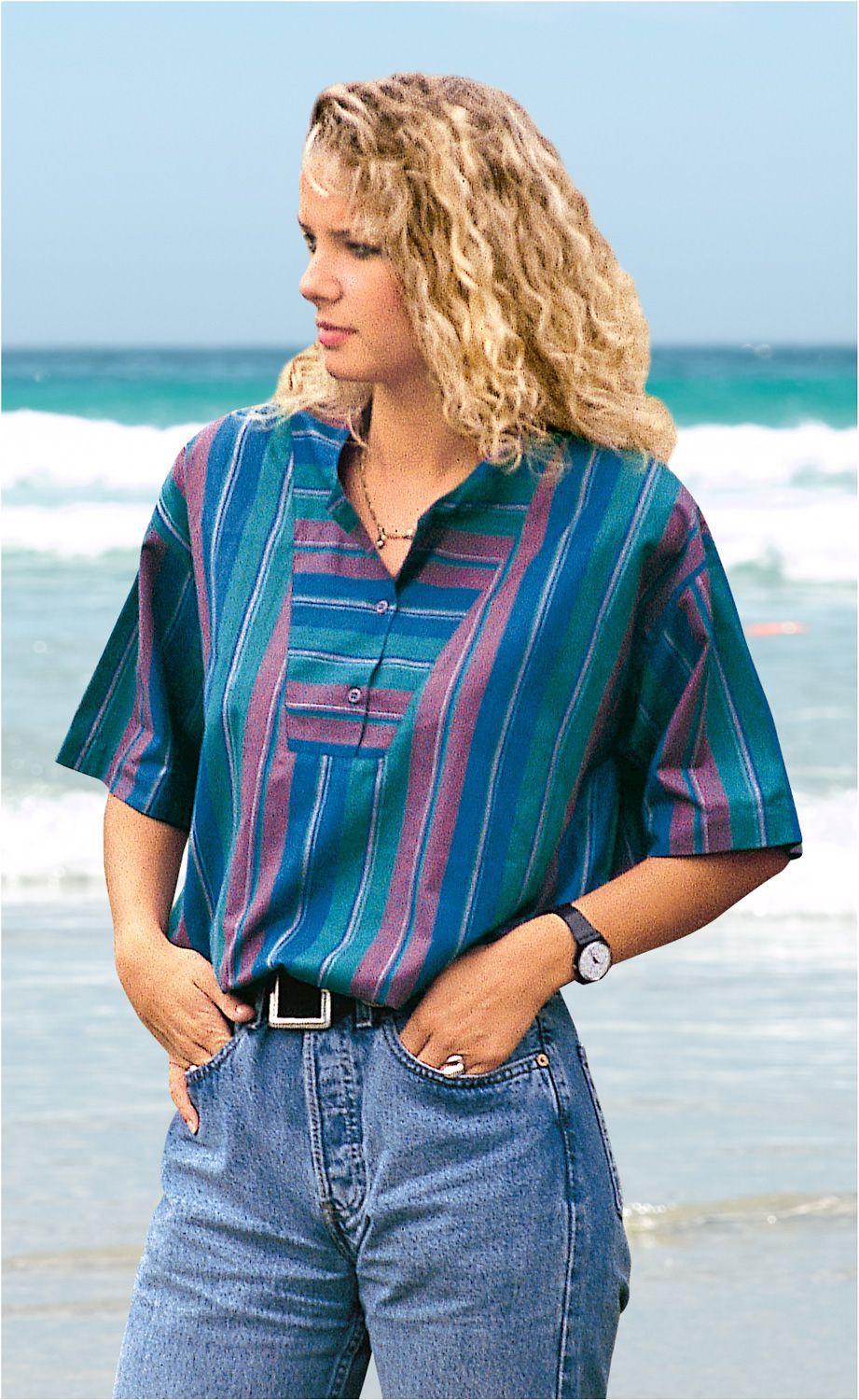 Frau am Strand trägt gestreifte Baumwoll-Bluse in Blau und Lila mit kurzem Arm, kombiniert mit blauer Jeans und schwarzem Gürtel, die Hände in den Taschen, lockige blonde Haare, Blick seitlich, blauer Himmel und Meer im Hintergrund