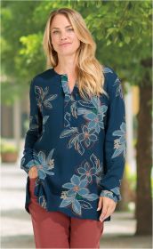 Frau mit langen blonden Haaren trägt eine dunkelblaue Modal-Bluse mit großem floralem Muster in hellblau, grün und beige, dazu rostrote Hose, steht vor einem grünen Hintergrund mit Bäumen und Weg