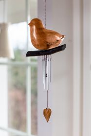 Windspiel Vogel aus hellem Holz in Vogelform, sitzend auf dunklem Holzast, mit vier silbernen Metallrohren und hängendem Holzherz, vor hellem Fensterhintergrund