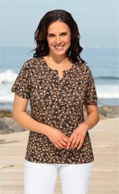 Frau trägt Viskose-Mix-Shirt mit schwarzem Grund und floralem Muster in Beige und Blau, steht am Strand auf Holzsteg, lächelt, dunkle gewellte Haare, weiße Hose, hellblauer Himmel und Meer im Hintergrund
