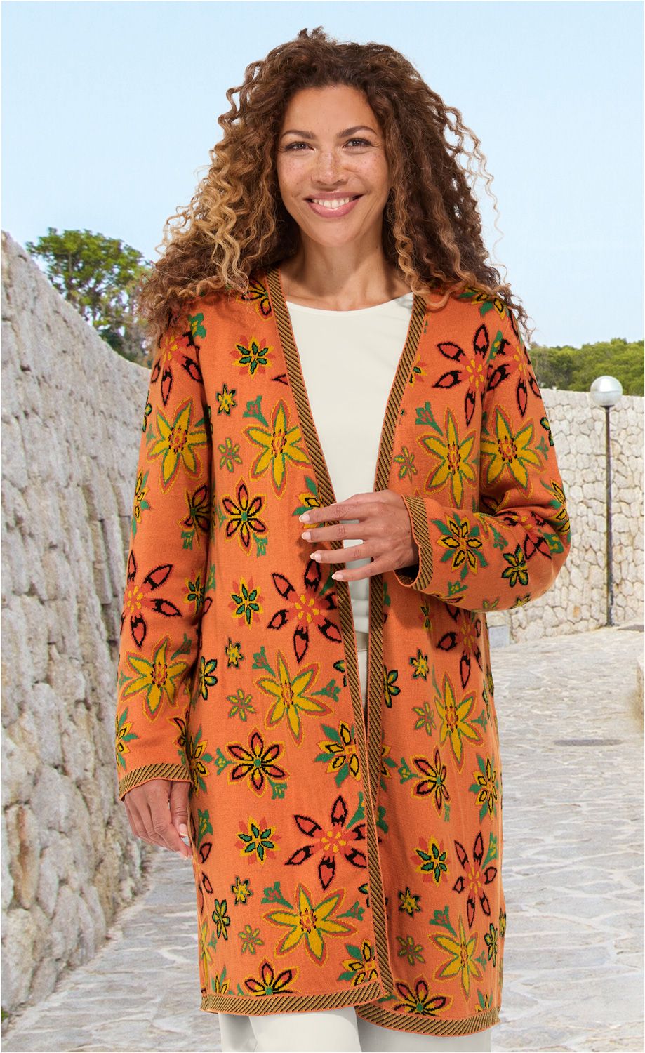 Frau trägt orangen Baumwoll-Mantel mit buntem Blumenmuster, offen getragen, vor hellem Outdoor-Hintergrund mit Steinmauern und gepflastertem Weg, lockige Haare, lächelnd